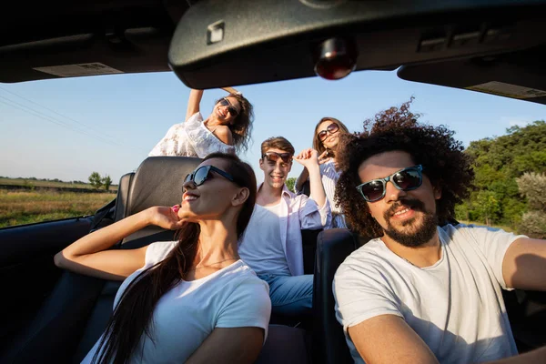 Stylish company of young girls and guys are sitting and smiling in a black cabriolet on the country road on a sunny day. — Stock Photo, Image