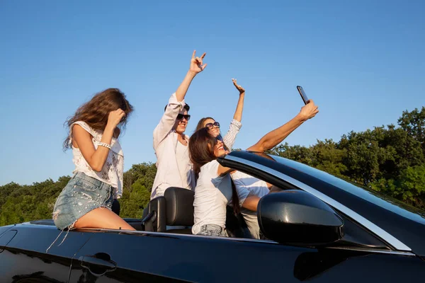 Joyeuses jeunes filles et les gars sont assis dans un cabriolet noir sur la route et faire du selfie par une journée ensoleillée . — Photo