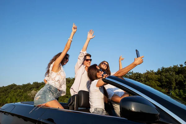 Joyeuses jeunes filles et les gars sont assis dans un cabriolet noir sur la route et faire du selfie par une journée ensoleillée . — Photo