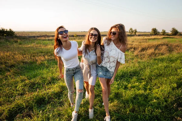 Drie prachtige jonge vrouwen in zonnebril gekleed in de mooie kleren staan in het veld en lachend op een zonnige dag. — Stockfoto