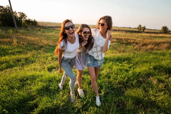 Tre bellissime giovani donne in occhiali da sole vestite con i bellissimi vestiti stanno sul campo e sorridono in una giornata di sole . — Foto Stock