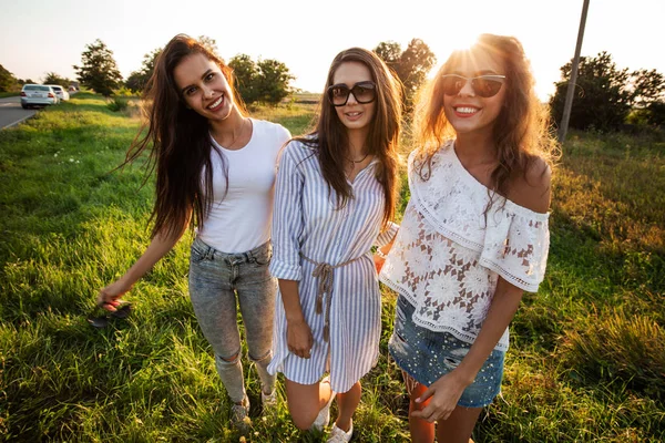 Τρεις πανέμορφες νεαρές γυναίκες σε γυαλιά ηλίου, ντυμένοι με όμορφα ρούχα της σταθεί στο πεδίο και χαμογελαστός σε μια ηλιόλουστη ημέρα. — Φωτογραφία Αρχείου