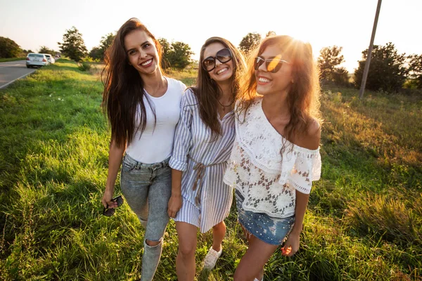 Tre bellissime giovani donne in occhiali da sole vestite con i bellissimi vestiti stanno sul campo e sorridono in una giornata di sole . — Foto Stock