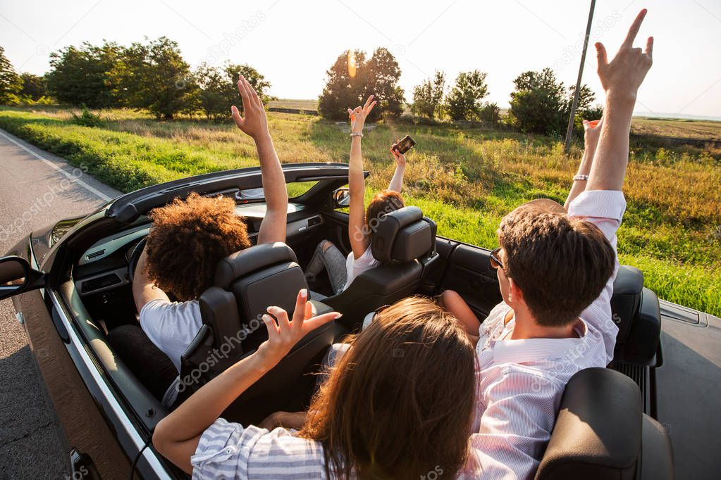 Young girls and guys are sitting in a black cabriolet, holding their hands up and making selfie on a warm sunny day.
