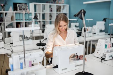 Bir akıllı görünümlü sarışın kadın beyaz gömlek giymiş ile elektrikli dikiş makinesi dikiş var. Moda, terziler atölye.