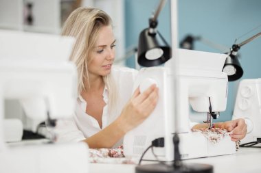 Bir akıllı görünümlü sarışın kadın beyaz gömlek giymiş ile elektrikli dikiş makinesi dikiş var. Moda, terziler atölye.