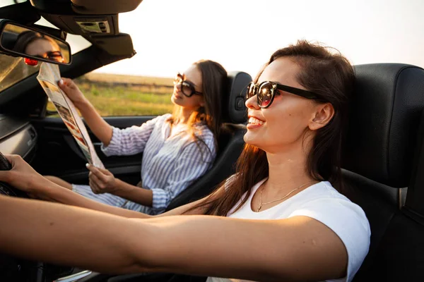 De belles jeunes femmes aux cheveux bruns en lunettes de soleil sont assises dans un cabriolet noir par une journée ensoleillée. L'un d'eux garde la carte entre ses mains. . — Photo