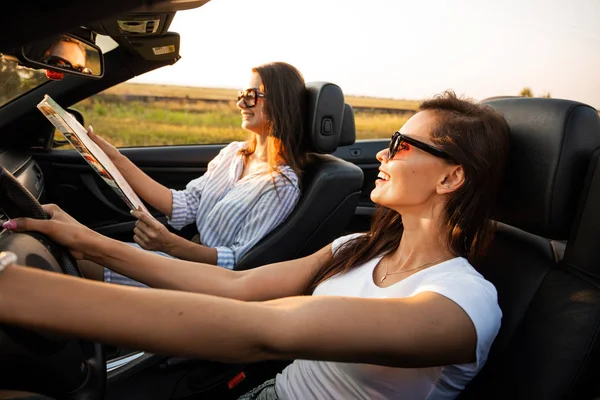 Beautiful dark-haired young women in sunglasses are sitting in a black cabriolet on a sunny day. One of them keeps map in her hands. — Stock Photo, Image