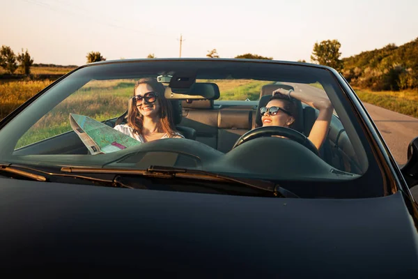 Belas mulheres jovens de cabelos escuros em óculos de sol estão sentadas em um cabriolet preto em um dia ensolarado. Um deles guarda o mapa nas mãos dela. . — Fotografia de Stock