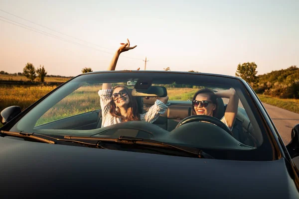 Two charming dark-haired young women in sunglasses are sitting in a black cabriolet on a sunny day. — Stock Photo, Image