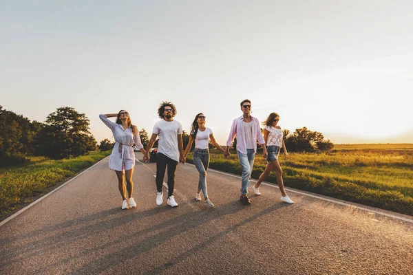 Compagnia di giovani ragazzi felici ed eleganti camminare su una strada di campagna in una giornata di sole — Foto Stock