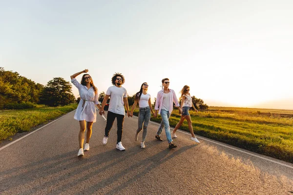 Empresa de jovens felizes e elegantes caminham em uma estrada rural em um dia ensolarado — Fotografia de Stock