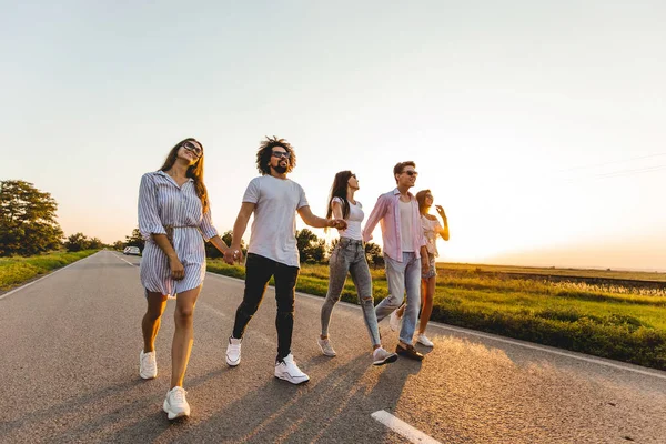 Gesellschaft fröhlicher junger stylischer Kerle hält sich an der Hand und geht an einem sonnigen Tag auf einer Landstraße spazieren — Stockfoto