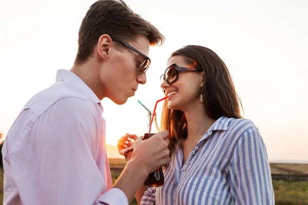 Schöne dunkelhaarige Mädchen und ein junger Mann mit Sonnenbrille trinken aus einer Flasche ein Getränk durch Strohhalme im Freien an einem sonnigen Tag. — Stockfoto