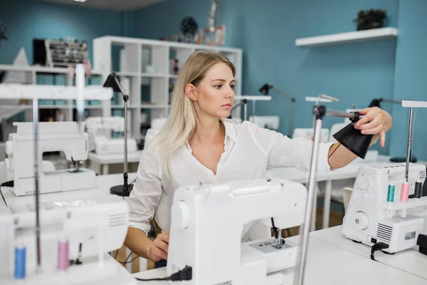Bir akıllı görünümlü sarışın kadın beyaz gömlek giymiş ile elektrikli dikiş makinesi dikiş var. Moda, terziler atölye.