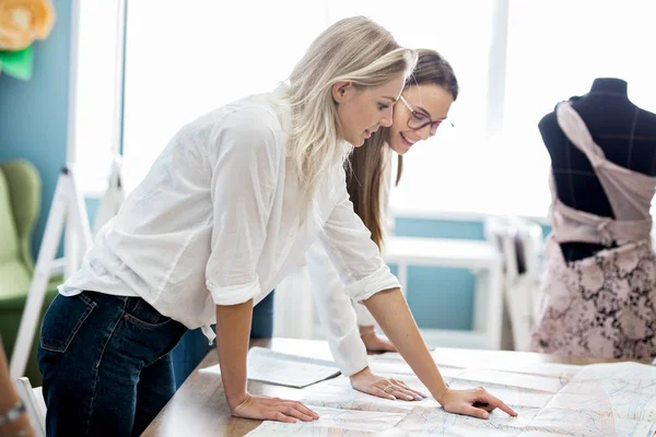 Dos mujeres guapas de aspecto inteligente están apuntando a la revista de costura. Moda, taller de sastres . — Foto de Stock