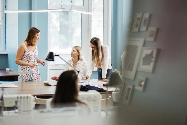 Tre kønne kvinder, der ser kloge ud, taler ved sybordet. Skrædderdukken er i baggrunden. Mode, skrædder workshop . - Stock-foto