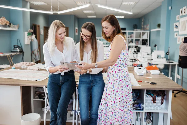 Trei femei frumoase cu aspect inteligent care poartă tricouri albe se uită la revista de cusut. Atelier de modă, croitori . — Fotografie, imagine de stoc