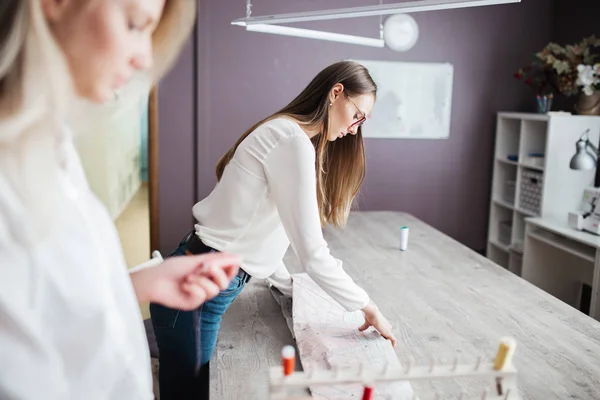 Twee smart uitziende mooie vrouwen het dragen van witte shirts zijn leunt over de tafel naaien. Mode, kleermakers workshop. — Stockfoto