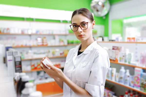 A young thin brown - haired lady with glasses, dressed in a white coat, looks at the camera and holds a small jar in the lobby of a new pharmacy.