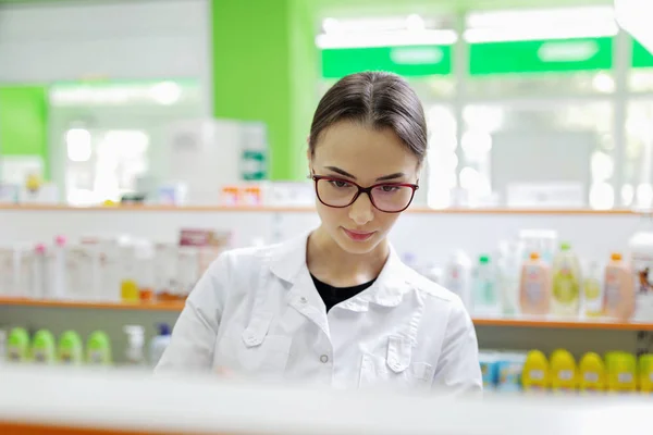 Una señora de pelo oscuro bastante elegante con gafas, vestida con un abrigo blanco, examina cuidadosamente los productos en el estante de la farmacia . — Foto de Stock