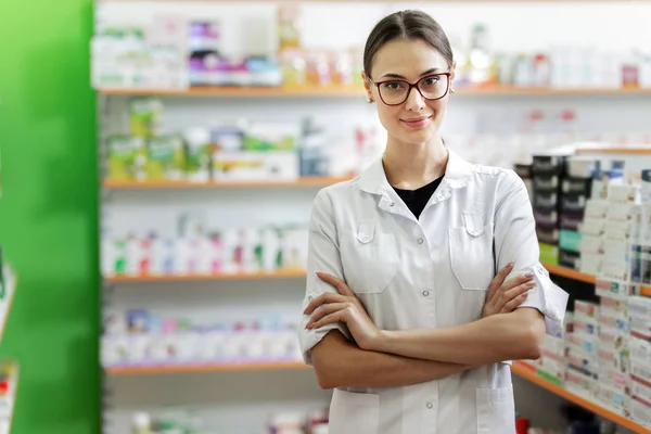 A smiling slim dark-haired lady with glasses, wearing a white coat, stands next to the shelf in the modern pharmacy.