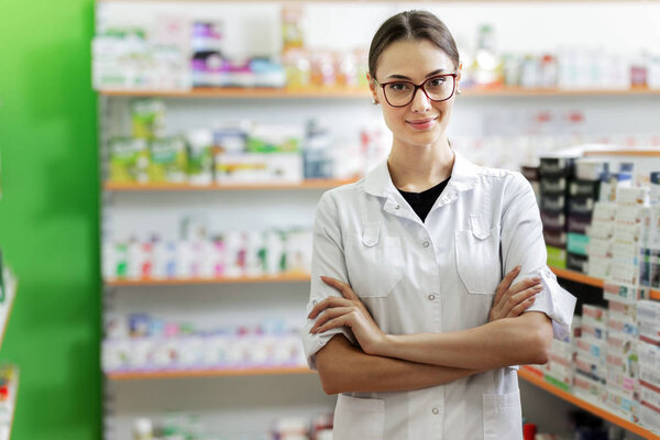 Улыбающаяся стройная темноволосая женщина в очках в белом халате стоит рядом с полкой в современной аптеке.
.