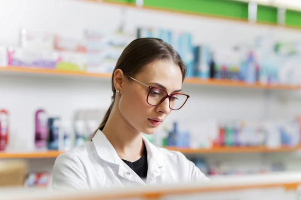 Una chica delgada de buen aspecto con cabello oscuro y gafas, con un abrigo blanco, coloca suavemente los productos en el estante en la farmacia . — Foto de Stock