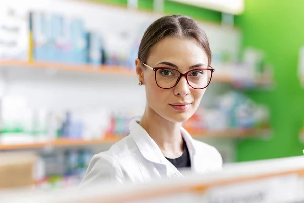 Una jovencita morena agradable y joven con gafas, vistiendo un uniforme médico, se para junto al estante en una farmacia moderna y mira a la cámara . — Foto de Stock