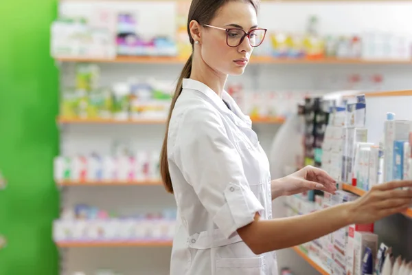 Una linda chica delgada con cabello oscuro y gafas, con una bata de laboratorio, se para junto al estante en una farmacia y lo examina cuidadosamente . — Foto de Stock