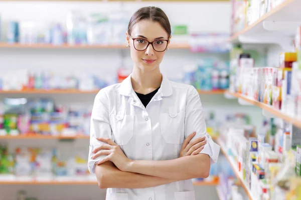Una joven delgada con el pelo largo y oscuro y gafas, con un abrigo blanco, con confianza se encuentra junto a los estantes en una farmacia actualizada . — Foto de Stock