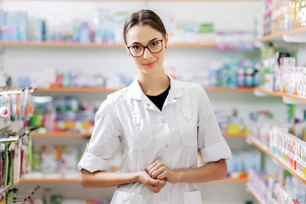 Una joven muy sonriente con el pelo oscuro y gafas, con un abrigo blanco, está junto a los sjelves en una farmacia con los brazos cruzados . — Foto de Stock