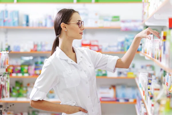 Una joven de cabello castaño delgado con gafas, vestida con una bata de laboratorio, toma algunos medicamentos del estante de una farmacia. El perfil de las niñas se muestra . —  Fotos de Stock