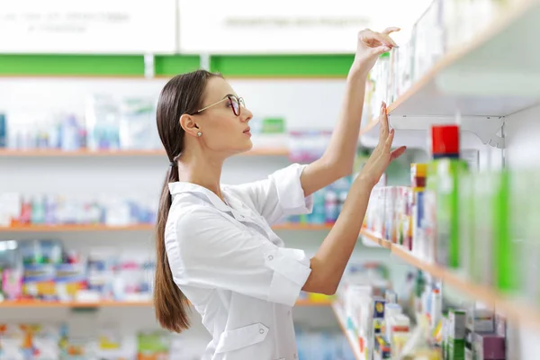 Uma menina bonita e magra com cabelo escuro e óculos, vestindo um casaco de laboratório, tira algo da prateleira em uma farmácia moderna . — Fotografia de Stock
