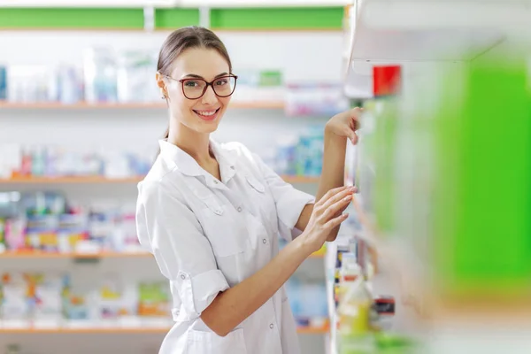A smiling slim dark-haired lady with glasses, wearing a white coat, stands next to the shelf in the modern pharmacy. Friendly atmosphere.