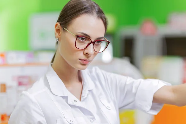 Una dama de pelo oscuro bastante elegante con gafas, vestida con una bata blanca, examina cuidadosamente los productos en el estante de la farmacia. . — Foto de Stock