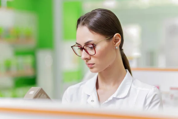 Una señora de pelo oscuro bastante elegante con gafas, vestida con una bata blanca, examina cuidadosamente los productos en el estante de la farmacia. Perfil de Girls se muestra . —  Fotos de Stock