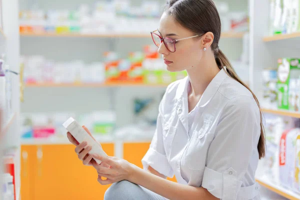 Una joven de cabello castaño delgado con gafas, vestida con un conjunto médico, lee algo en el paquete de pastillas junto a los estantes de la farmacia . — Foto de Stock