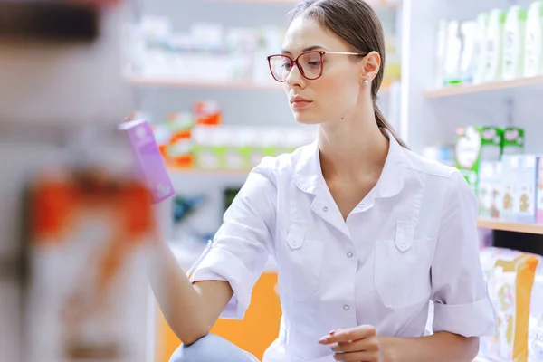 Una joven de cabello castaño delgado con gafas, vestida con una bata de laboratorio, agachada, examina los medicamentos en el estante de una farmacia . — Foto de Stock