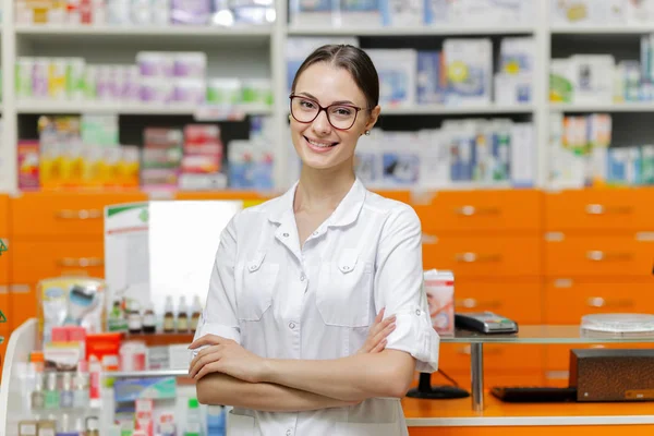 Una bella ragazza sorridente con i capelli scuri e gli occhiali, con indosso un complesso medico, sta vicino alla cassa con le braccia incrociate in una farmacia moderna. . — Foto Stock