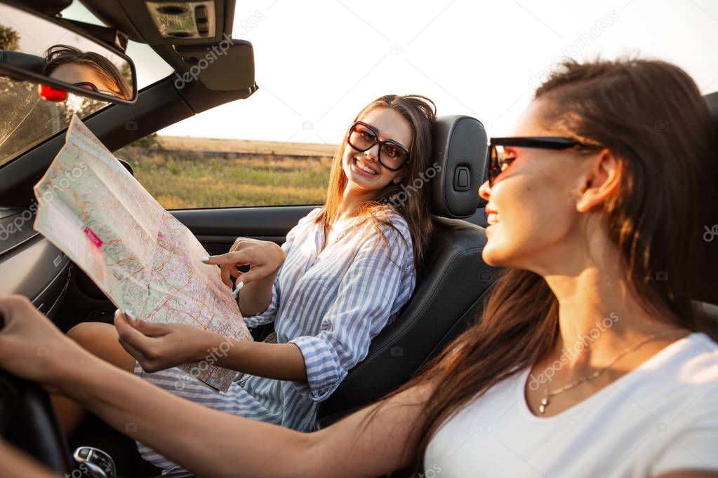 Beautiful dark-haired young women in sunglasses are sitting in a black cabriolet on a sunny day. One of them keeps map in her hands.