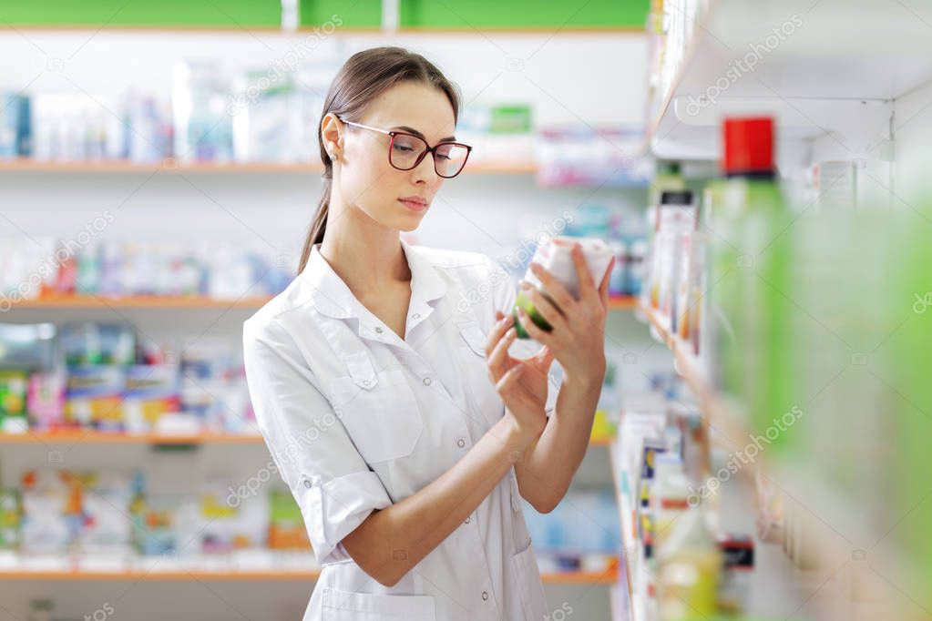 A young thin brown-haired girl with glasses, dressed in a medical overall, reads something on the pills pack next to the shelves in the pharmacy.