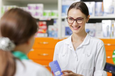 A cute slim girl with long dark hair and glasses,wearing a white coat,is talking with a visitor in a new stylish pharmacy. . clipart
