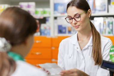 A friendly slim lady with  dark hair and glasses,wearing a lab coat,is talking with a visitor and reads a prescription in a new stylish pharmacy. clipart