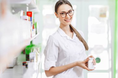 Beyaz bir önlük giyen bir gülümseyen sevimli ince esmer kadınla gözlüklü, raf yanında duruyor ve küçük bir kavanoza modern eczanede gösterir. Onun ince profiliyle gösterilir.