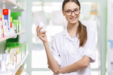 A youthful thin brown-haired lady with glasses,dressed in a lab coat,is holding a small jar in her right hand in a new pharmacy, clipart