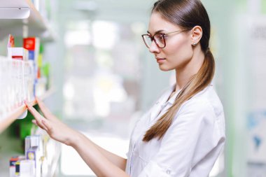 Genel olarak, bir tıbbi giyen oldukça ince kız siyah saçlı ve gözlük, ile raf bir şey yeni bir eczane alır.