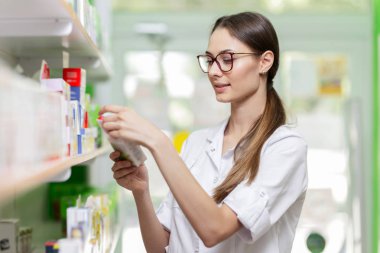 Bir laboratuar önlüğü giymiş bir büyüleyici gülümseyen kız siyah saçlı ve gözlük, raf bir şey modern bir eczane alır.