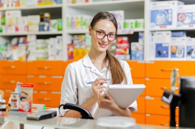 Güzel bir ince esmer kız gözlüklü bir laboratuar önlüğü giymiş bir defter modern bir eczane nakit masasında notlar yazıyor.