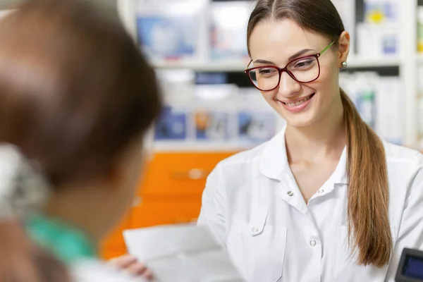 Una dama delgada sonriente con cabello oscuro y gafas, con una bata de laboratorio, está hablando con un visitante y lee una receta en una farmacia actualizada . —  Fotos de Stock